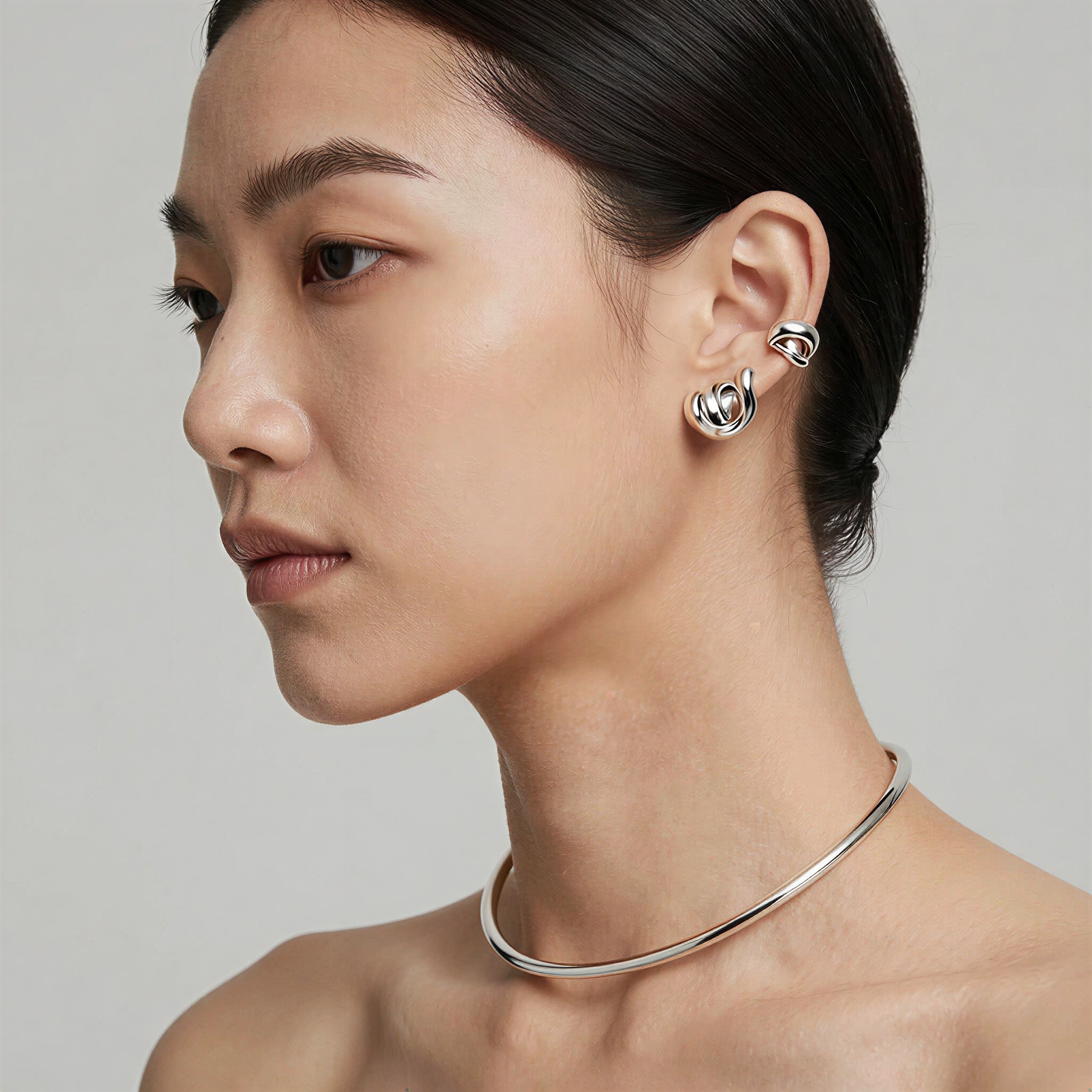 A close fashion-beauty portrait focused on an adult model wearing a sculptural silver ear cuff and matching collar necklace. Her hair is pulled back cleanly, ma